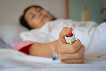 Woman patient  receiving intravenous fluid through IV line holding red emergency call button while lying in hospital bed.Hand holding  nurse call button,selective focused.の写真素材