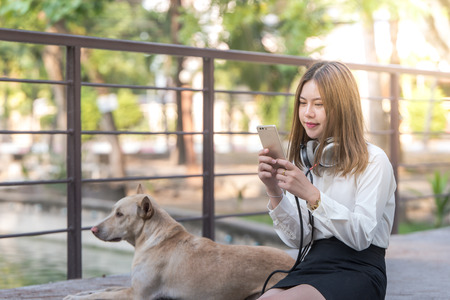 Portrait of a businesswoman young happy girl listening music on line with headphones from a smartphone on the bridge in the park outdoor summer sunny day .relax with her dogの写真素材
