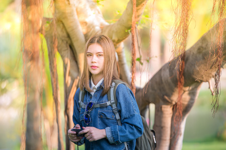 Outdoor fashion portrait traveler of stylish photographer girl holding camera, wearing jeans jacket, trendy sunglasses and backpack, amazing view of forest with sunsetの写真素材