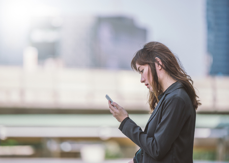 beautiful young woman is using an app in her smartphone device to send a text message in front of a city background she wait text from boyfriendの写真素材