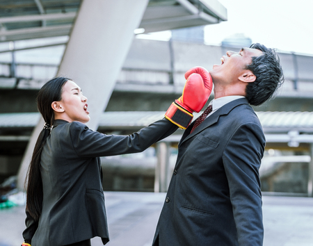 Business concept : Young Asian business woman in suit uniform wearing boxing gloves and punching business man's faceの写真素材