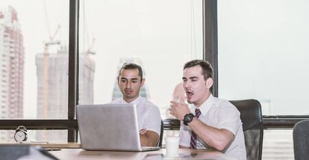 Young yawning businessman working with laptop at desk in the modern office, unmotivated employee, not completing job duties, do not have the drive to succeed, dissatisfaction in office environmentの写真素材