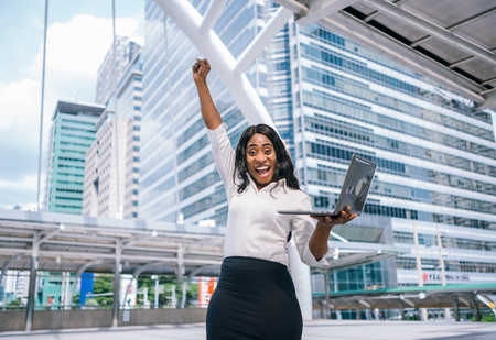 Cheerful African American business woman with arms raised in success and hold laptopの写真素材