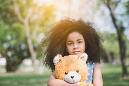 Little cute african american girl hugging embraces an amusing teddy bear against summer nature.の写真素材