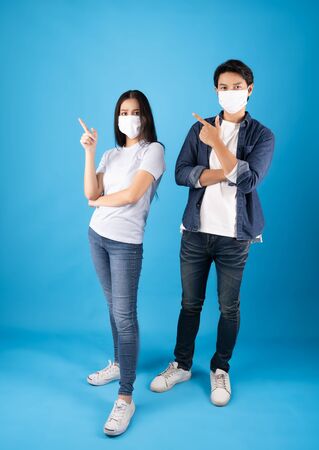 Young happy Asian man and woman pointing hands up to copy space wearing hygienic mask to prevent infection corona virus Air pollution pm2.5  shoot on blue background looking at cameraの写真素材