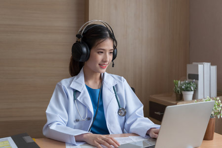 Asian Female doctor wear headphones working at office desk and smiling at camera, video call consultation, patient and doctor talk concept.の写真素材