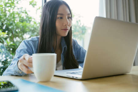 Asian woman using a laptop working at home hold a coffee cup while sitting at work.の写真素材