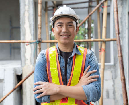 Portrait of Asian man engineer at building site looking at camera. Male construction manager wearing white helmet and yellow safety vestの写真素材