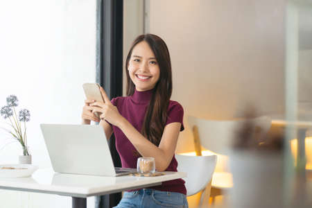 Young Asian woman using phone and laptop in coffee shop and looking at camera.の写真素材