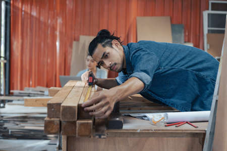 Young Asian man Carpenter uses a tape measure to measure wood on the workbench in woodcraft carpentry workshop.の写真素材