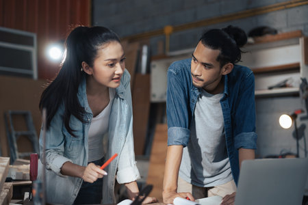 Young Asian couple carpenters man and woman discussing about design of products on laptop in woodcraft carpentry workshop.の写真素材