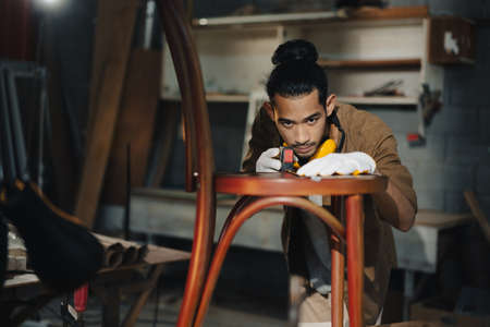 Young Asian man Carpenter uses a tape measure to measure chair on the workbench in woodcraft...