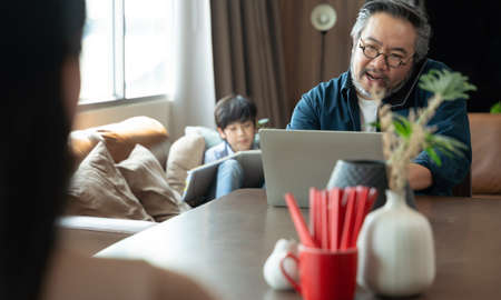 Asian middle aged man sitting at home working with family at home. Work from home conceptの写真素材