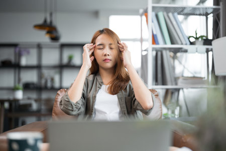 Young Asian woman overworked and exhausted experiences headaches and vision issues after using a laptop for work.の写真素材