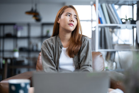 Young Asian woman working on laptop computer looking away thinkingの写真素材