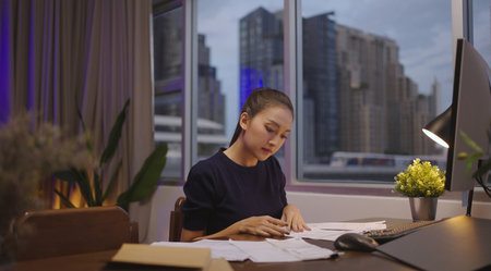 Asian woman sits at her desk with paperwork and computer at home working late at night.の写真素材