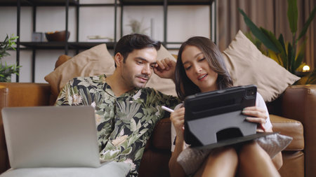 Young couple using tablet and laptop together on sofa in living room at homeの写真素材