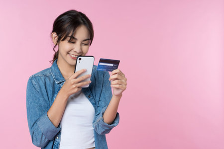 Young beauty Asian woman shopping payment online with credit card on smartphone and she wearing jean and looking at phone isolated on pink backgroundの写真素材
