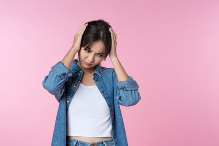 Young Asian woman is suffering from a severe headache, pressing her hand to her temples and frowning to relieve pain, with a helpless expression on her face.の写真素材