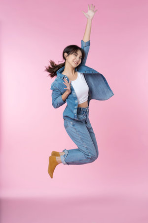 Elegant young Asian woman raises her arm and leg in a studio shot, expressing happiness and success isolated pink backgroundの写真素材