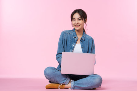Young Asian college student sits cross-legged on floor working with a laptop computer, looking directly at the camera in a studio shot on a pink backgroundの写真素材