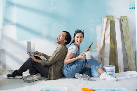 Young Asian couple repairing and painting the wall Sit and look at home decorating ideas through a laptop.の写真素材