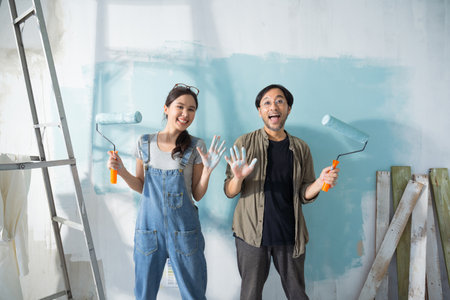 Young Asian couple repairing and painting the wall with blue paint using a roller during renovation in their new apartment.の写真素材