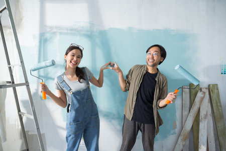 Asian couple expressing love heart while painting the wall with light blue paint using a roller during renovation in their new apartment.の写真素材