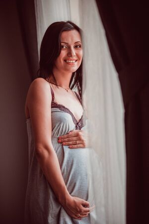 Pregnant woman put her hands her belly on standing at the window