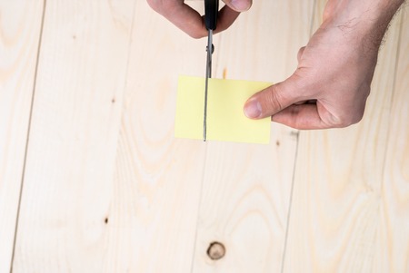 A man is cutting a sheet of yellow paper using metallic scissors on wooden background