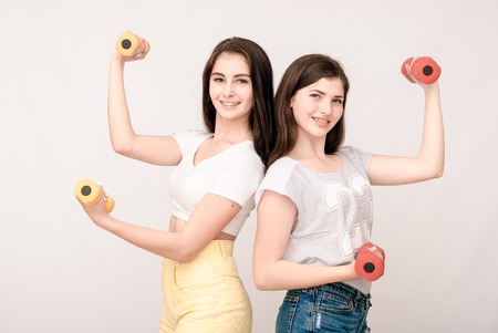 Positive friends portrait of two happy sister girls with dumbbell, funny faces, grimaces, joy, emotions, casual style on light grey backgroundの写真素材