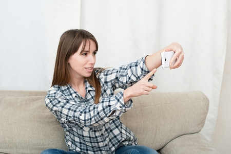 Young beautiful woman using her smartphone on the couch at home in the living room. Cute blond girl chating with friend or shopping onlineの写真素材