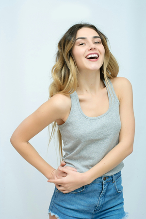 Young girl with long curly hair posing on blue background in studio. She wears shorts, pink T-shirt, pink sunglasses. She keeps hat overhead, holds blue skateboard on booty, looks excited.の写真素材