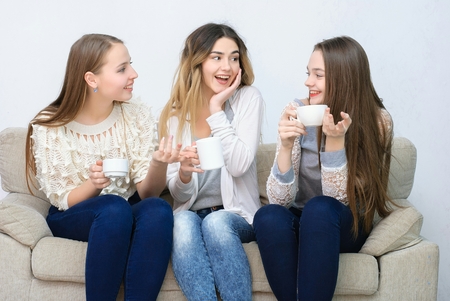 Three happy friends talking and drinking coffee and tea sitting on a couch at homeの写真素材