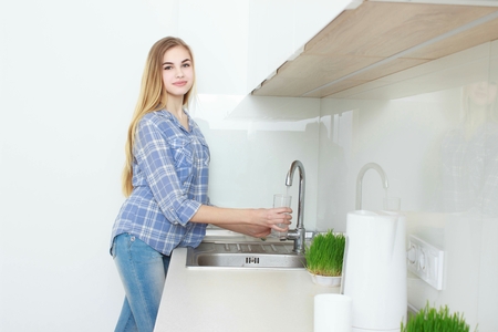Young beautiful girl in a blue checkered shirt and jeans in the kitchen pouring the glass of water. Morning. home interior.の写真素材