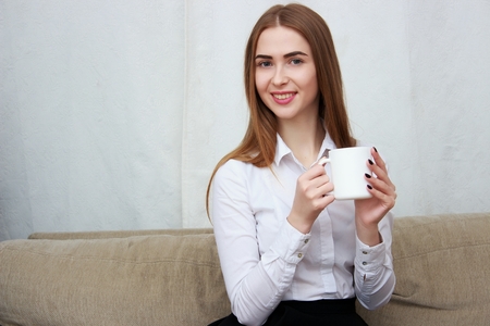 Young beautiful girl with cup of coffee sitting on couch at home.の写真素材