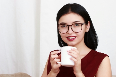 Beautiful girl in red sweater sitting on couch posing and smiling with cupの写真素材