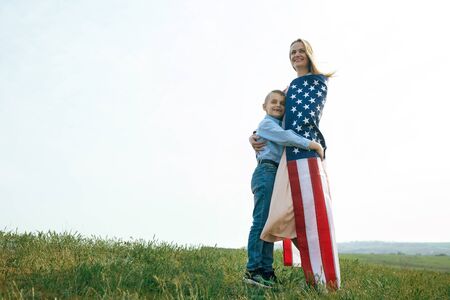 single mother with son on independence day of USA. Woman and her child walk with the USA flag on the ocean coast. Wonam dressed in a coral dress with a denim jacket.の写真素材