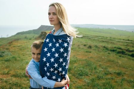 single mother with son on independence day of USA. Woman and her child walk with the USA flag on the ocean coast. Wonam dressed in a coral dress with a denim jacket.の写真素材