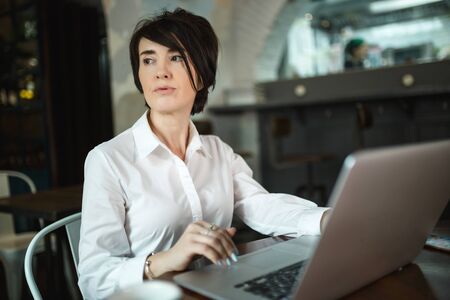 A middle-aged woman sits in a cafe, drinks coffee and works at a computer. A woman is thinking about somethingの写真素材