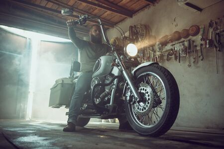 Handsome brutal man with a beard sitting on a motorcycle in his garage.