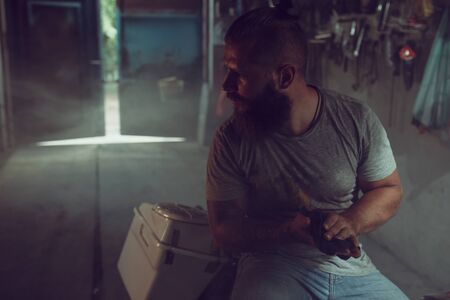 Handsome brutal man with a beard sitting on a motorcycle in his garage, wiping his hands and looking...