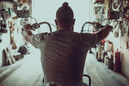 Handsome brutal man with a beard sitting on a motorcycle in his garage, view from the back.