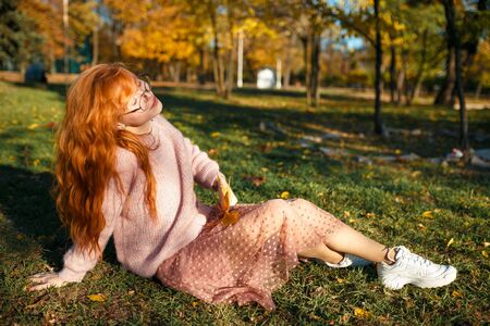 Portraits of a charming red-haired girl with a cute face. Girl posing in autumn park in a sweater and a coral-colored skirt. The girl has a wonderful mood.の写真素材