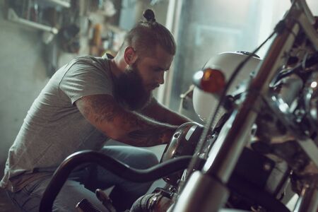 Handsome bearded man repairing his motorcycle in the garage. A man wearing jeans and a t-shirt.の写真素材