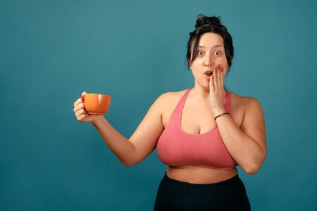 Happy plus size positive woman. Happy body positive concept. I love my body. Attractive overweight woman posing on camera in the studio with tea cup in her hands. Girl is wearing a coral braの写真素材
