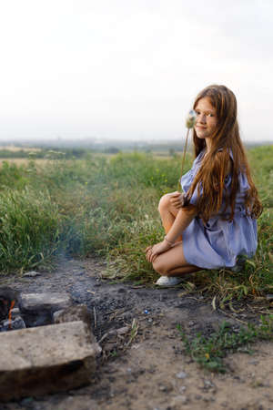 charming girl in a blue dress fries marshmallows in natureの写真素材