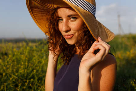 wonderful red-haired curly girl enjoys wonderful weather in nature. She is wearing a dress and a straw hatの写真素材