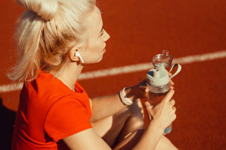 young beautiful blonde sits on a jogging track with a bottle of water in her hands