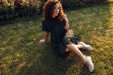 adorable curly red-haired girl with freckles in a dress enjoys good weather sitting on the lawnの写真素材